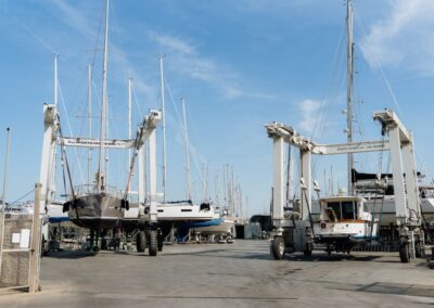 Due barche sollevate da gru mobili in un cantiere navale, circondate da altre imbarcazioni a vela