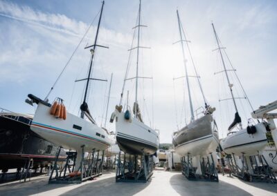 Barche a vela fuori dall’acqua sostenute da cavalletti in un cantiere navale, con cielo soleggiato sullo sfondo
