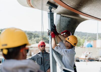 Operatori con caschi da cantiere posizionano un supporto sotto la chiglia di una barca sollevata